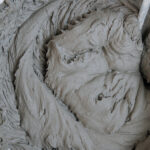 A bowl of grayish brown substance is being stirred with a spoon. The mixture appears to be a type of cement or concrete